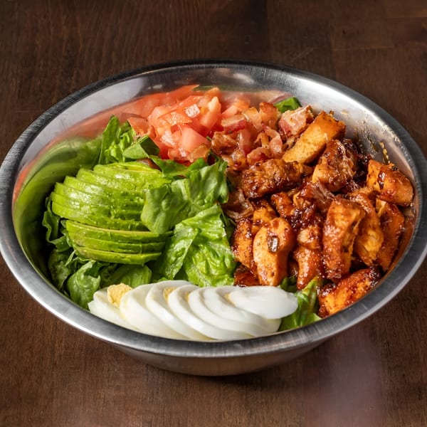 A large metal bowl filled with a salad of lettuce, avocado, tomatoes, bacon, chicken, and sliced eggs.