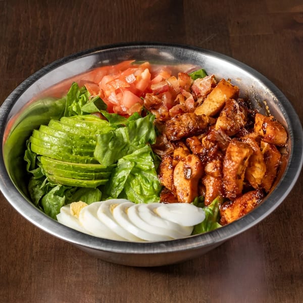 A large salad with lettuce, sliced avocado, diced tomatoes, bacon bits, sliced hard-boiled eggs, and grilled chicken.