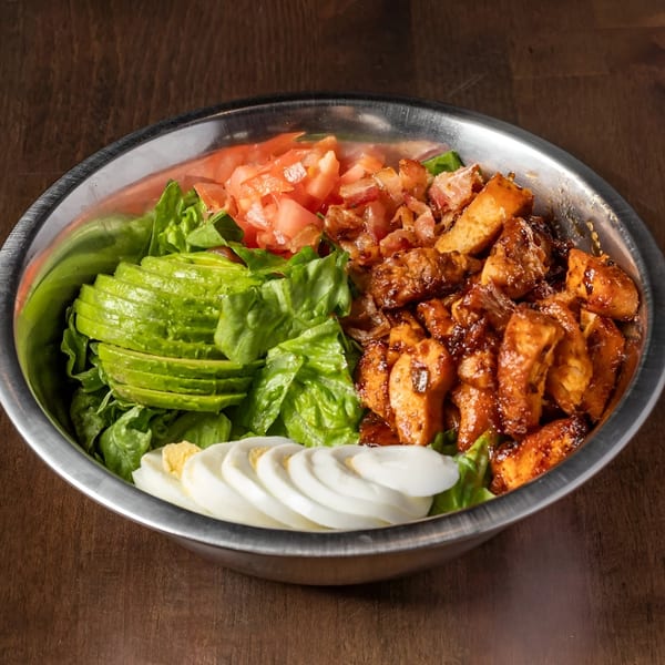 A large metal bowl filled with a salad featuring lettuce, avocado, tomatoes, bacon, chicken, and sliced hard-boiled eggs.