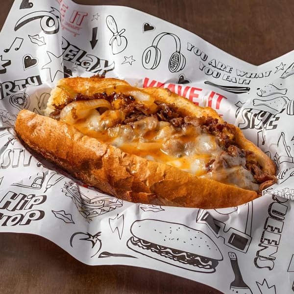 A Krave It cheesesteak sandwich with melted cheese and onions on a hero roll, in branded paper.