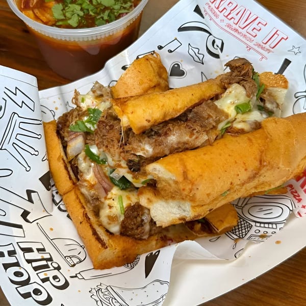 A Krave It cheesesteak sandwich with melted cheese and onions, next to a cup of dipping sauce.